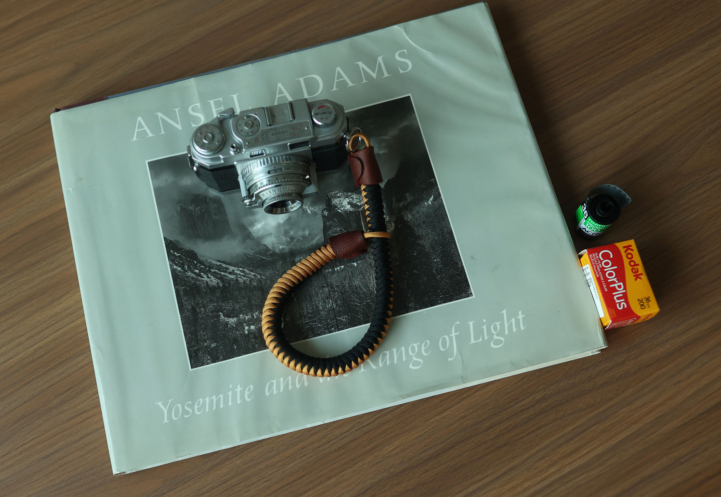 Black and gold braided camera wrist strap attached to a vintage film camera beside an open book