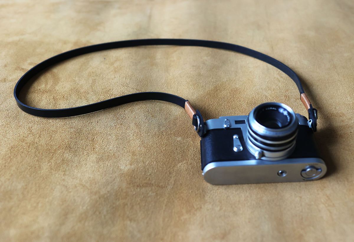 Minimalist black leather camera strap shown with a film camera on a wooden surface